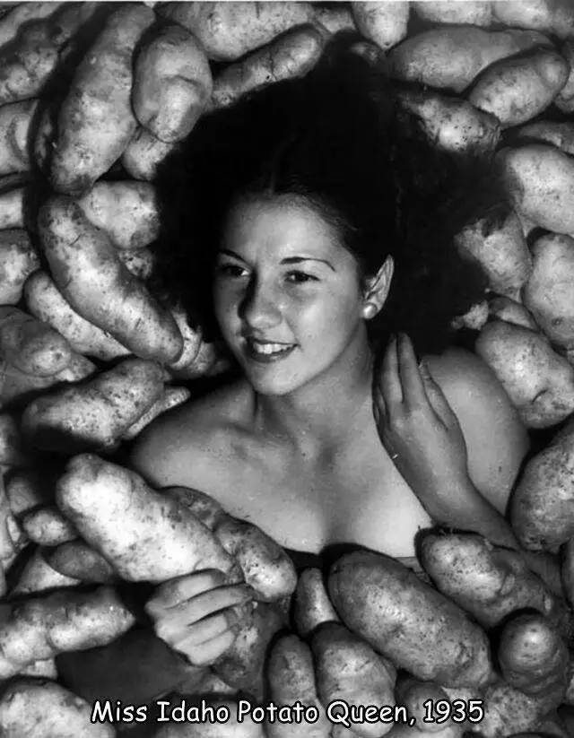 A vintage black-and-white photograph shows a smiling young woman with dark hair surrounded by a large pile of potatoes, with…