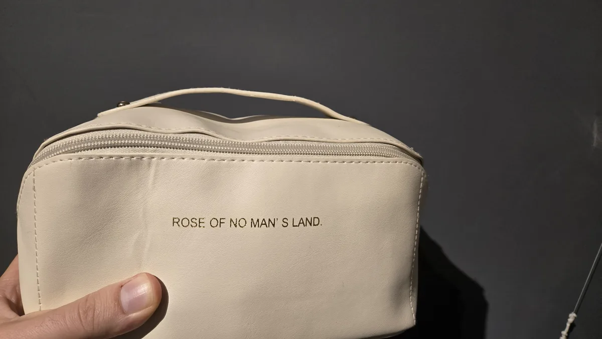 A hand holds a beige, zippered cosmetic bag against a dark gray background. The bag has gold text embossed on its front that…