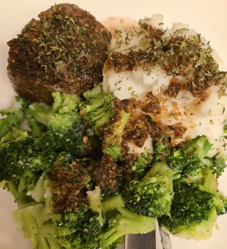 A close-up photo of a plate with a meatloaf patty, mashed potatoes, and steamed broccoli, all generously covered in a brown…