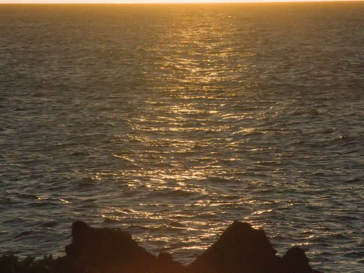 A serene sunset over the ocean with golden light reflecting on the water, silhouetted rocks in the foreground. The overlaid…