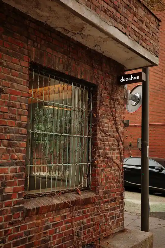A weathered brick building exterior features a window with ornate green metal bars and dried vines climbing the wall. A…