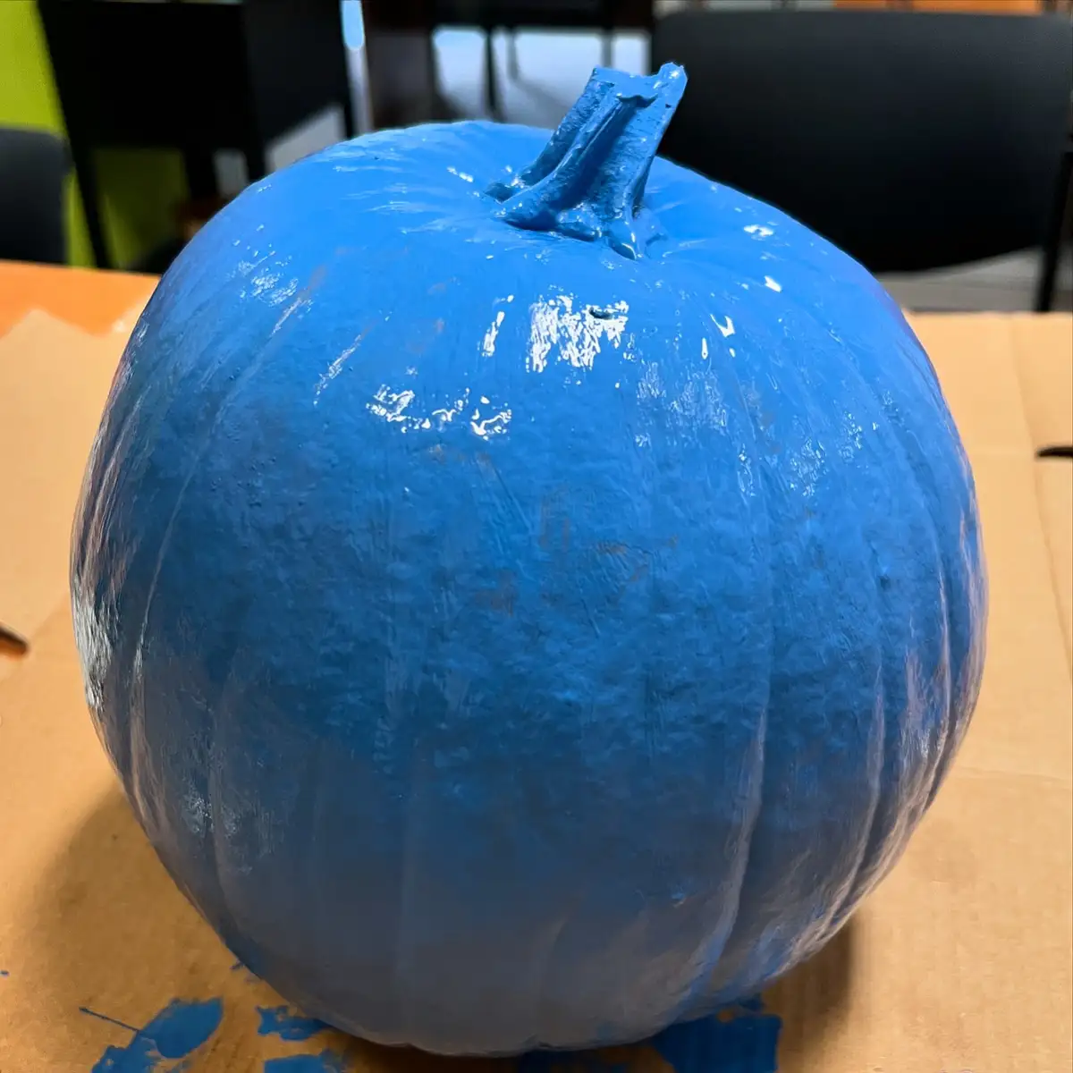 A close-up photo shows a pumpkin entirely covered in bright blue paint, sitting on a cardboard surface with paint splatters…