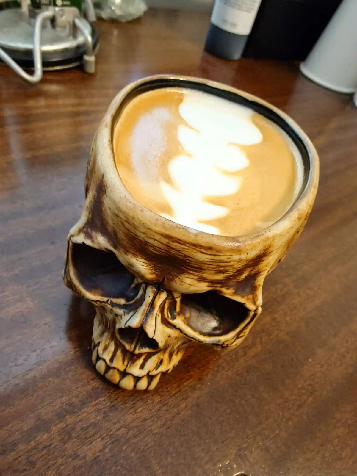 A coffee drink with latte art sits inside a ceramic mug shaped like a human skull, placed on a wooden table. The skull mug…
