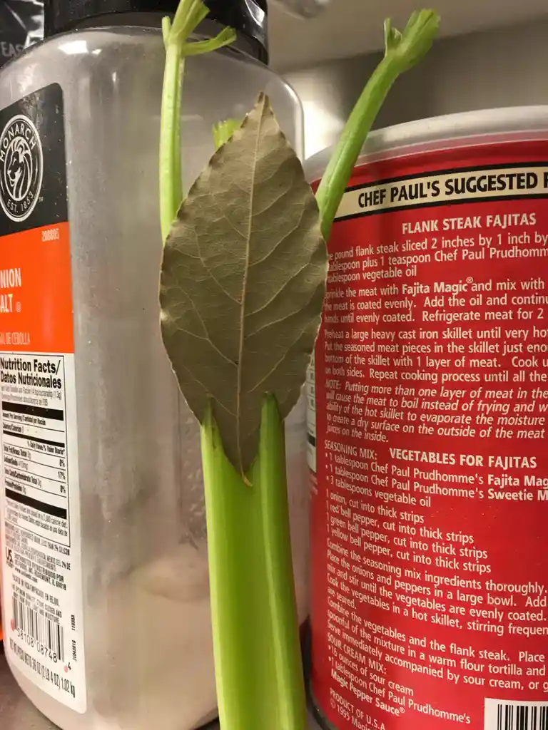 A close-up photo shows a celery stalk with a bay leaf stuck to it, positioned between a jar of onion salt and a red can of…