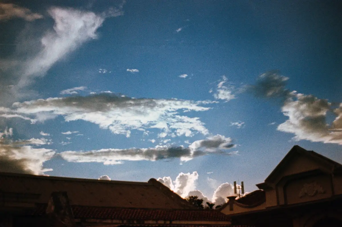 A low-angle shot shows the silhouette of tiled rooftops against a bright blue sky with scattered white clouds, some…