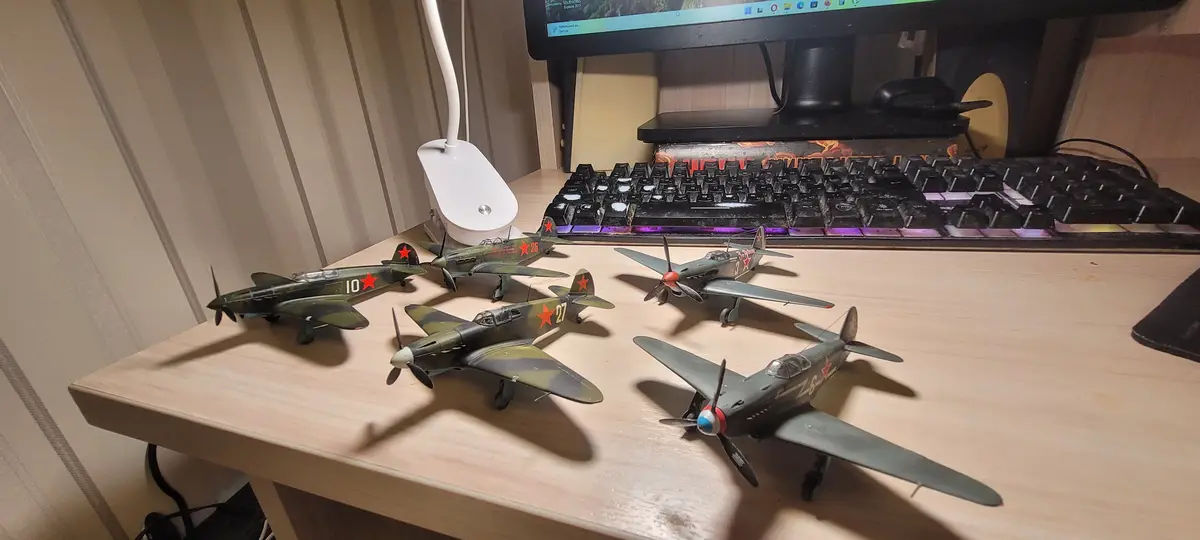 Five model airplanes with red star markings are arranged on a wooden desk in front of a computer setup with a keyboard and…