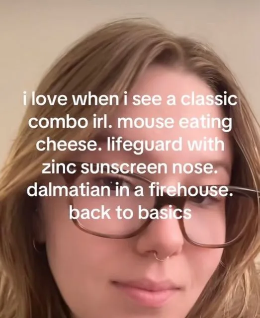 A close-up selfie of a young woman with light brown hair, wearing glasses and a nose ring, looking at the camera with a…