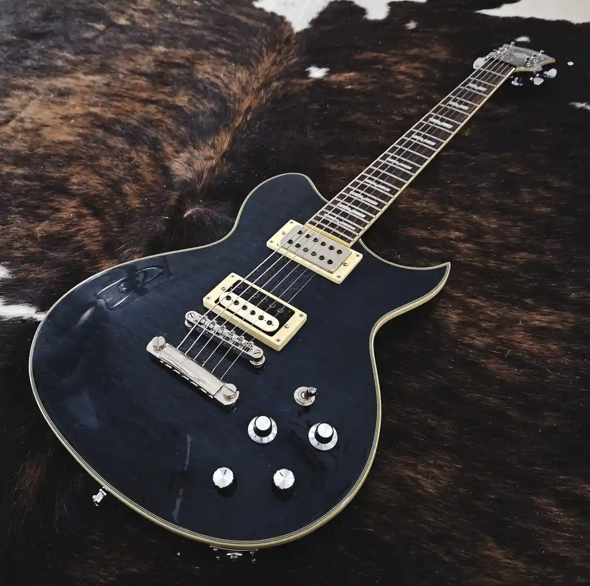 A black electric guitar with gold hardware rests diagonally on a brown and white cowhide rug. The guitar has two humbucker…