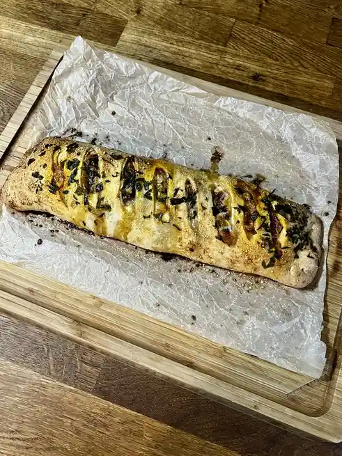 A long, baked savory bread loaf rests on parchment paper atop a wooden cutting board, its surface scored with diagonal cuts…