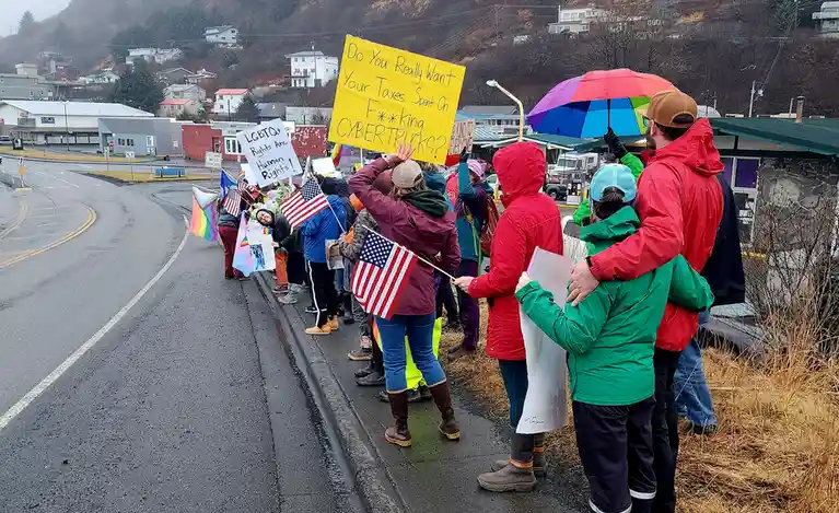 A group of protesters in raincoats and boots stand along a wet road holding signs and flags, including a large yellow sign…