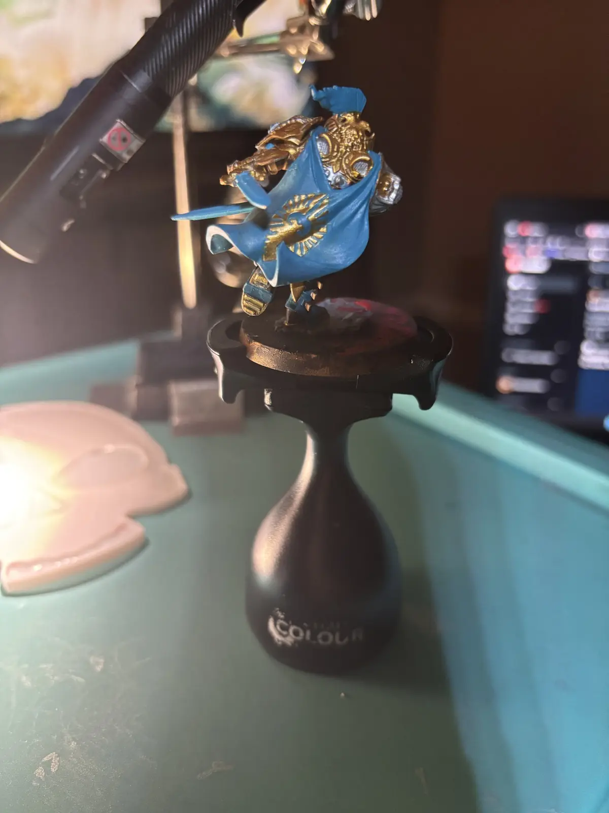 A painted miniature figurine of a knight in blue and gold armor, standing on a black 'Games Workshop Colour' stand, with a…