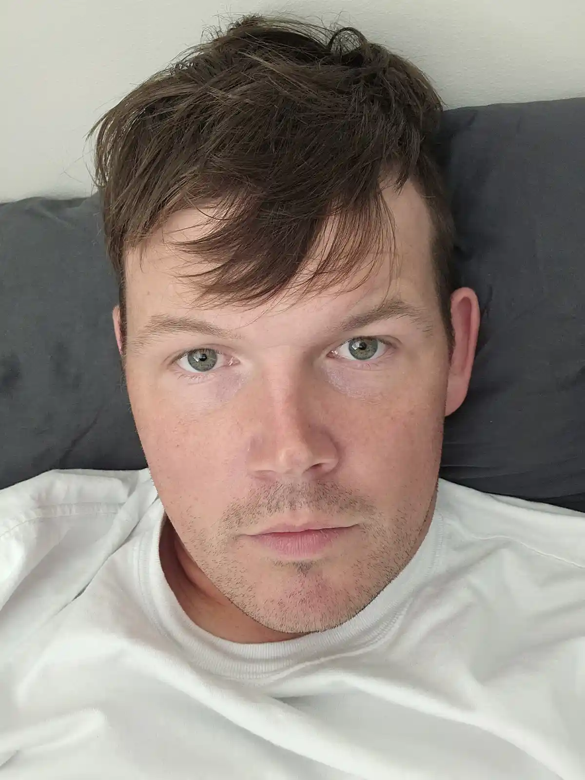 A man with brown hair and green eyes lies in bed on a gray pillow, wearing a white t-shirt, staring directly at the camera…
