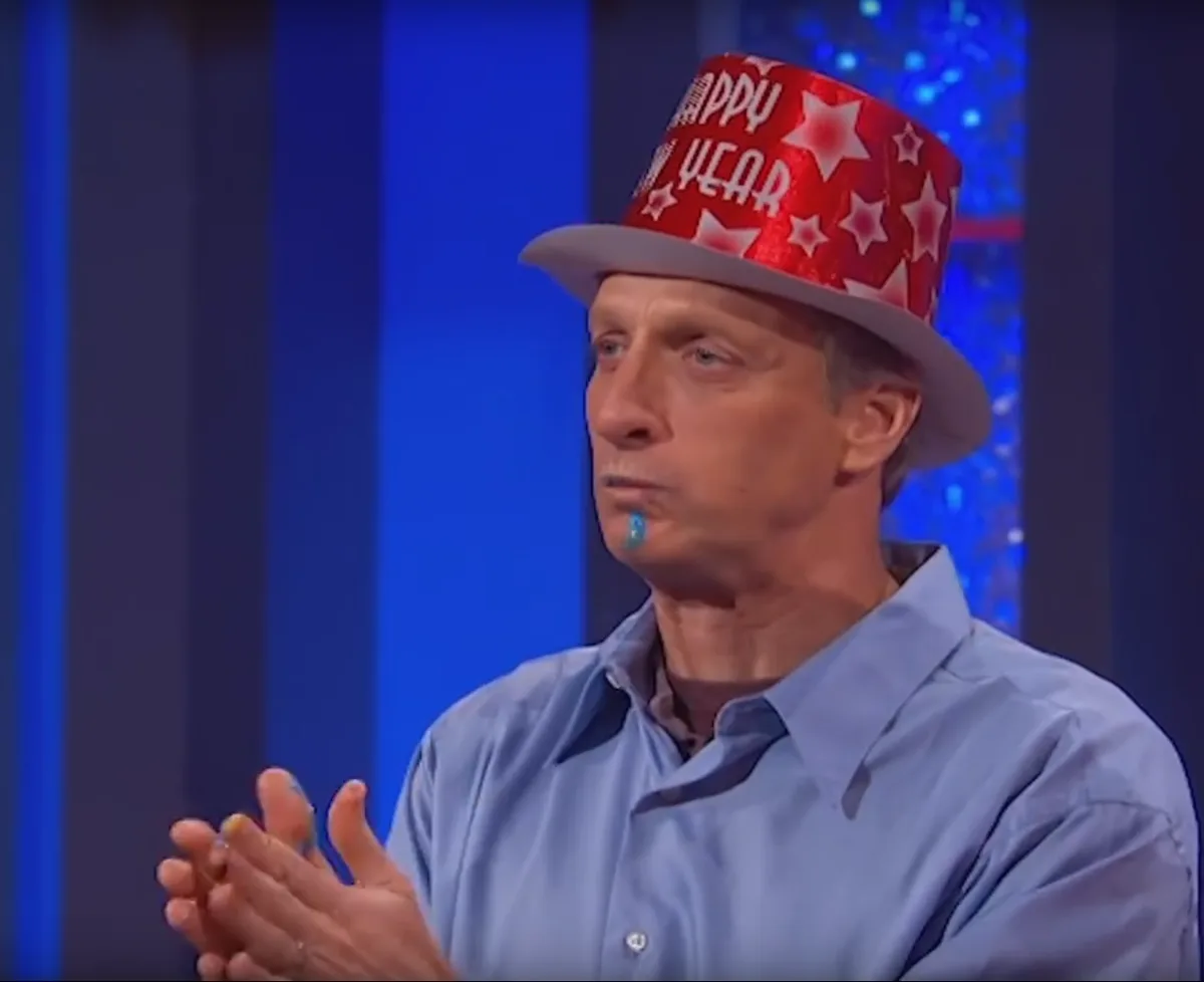a man wearing a red and white 'happy new year' party hat with stars, has blue confetti stuck to his chin and is clapping his…