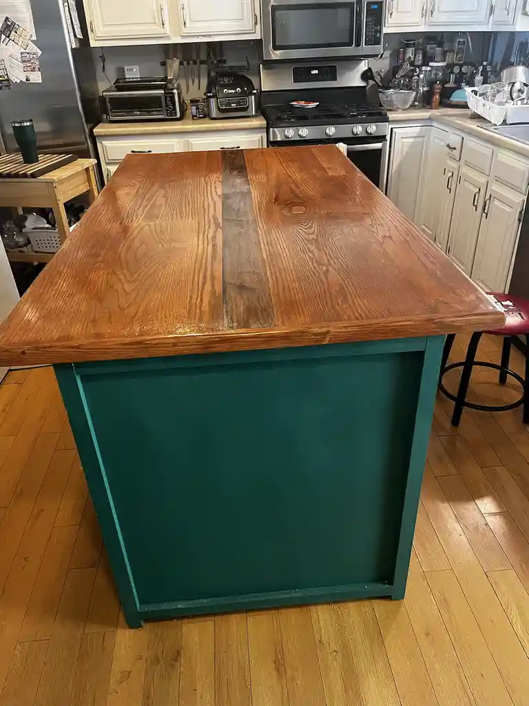 A photo of a kitchen island with a wooden top and teal-painted base, set in a home kitchen with white cabinets and stainless…
