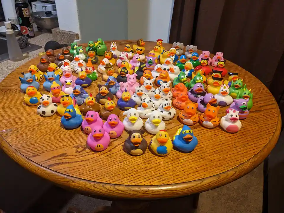 A large wooden table is covered with dozens of colorful, customized rubber ducks, each uniquely painted or decorated with…