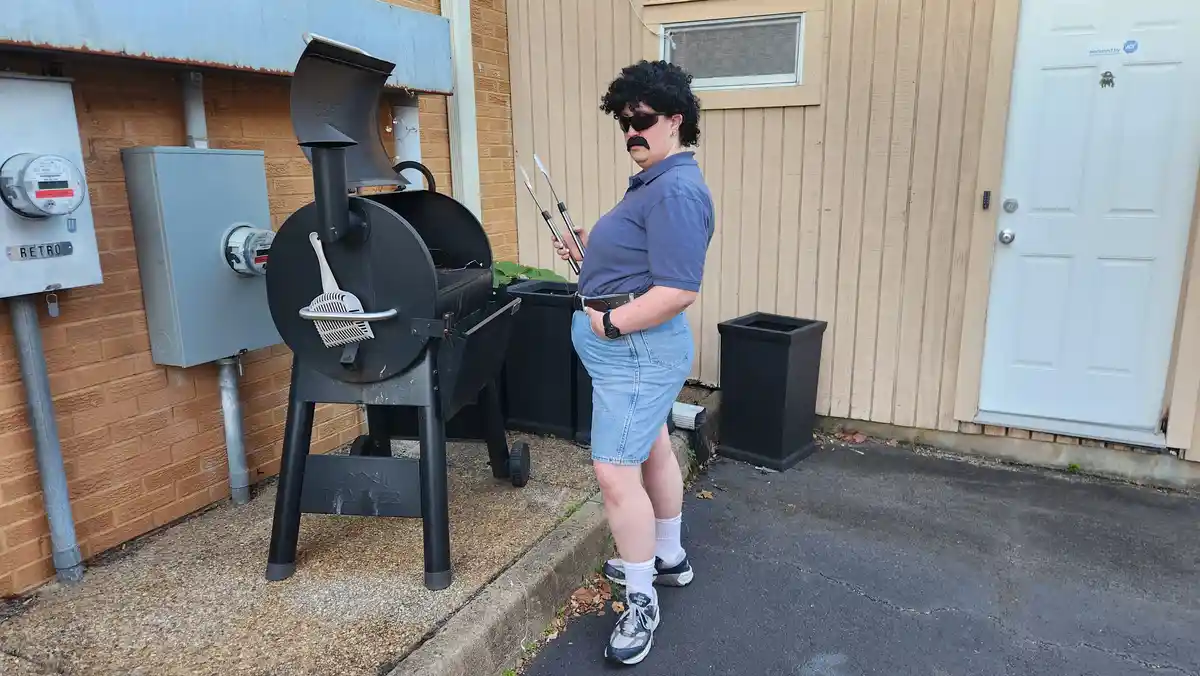 A person wearing a black curly wig, sunglasses, and a fake mustache stands next to a large black Traeger grill, holding…
