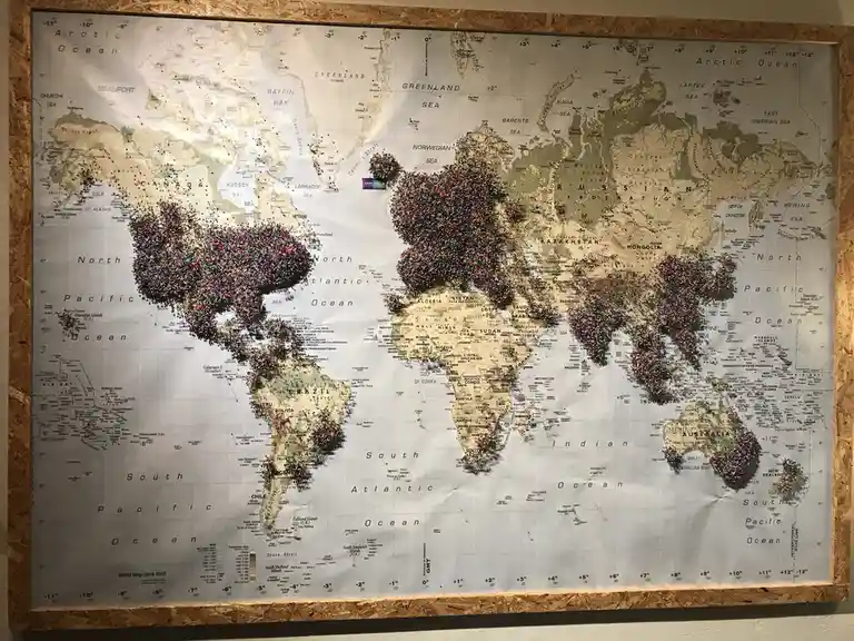 A framed world map from June 2010 is densely covered with thousands of colorful pushpins marking visited locations, with the…