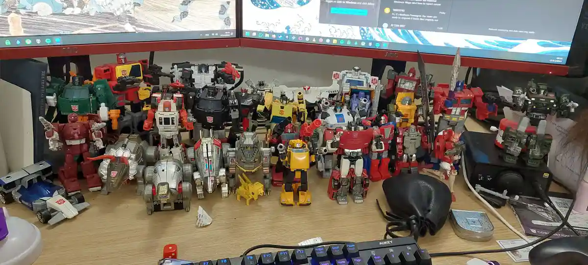 A wooden desk is covered with dozens of Transformers action figures in various sizes, colors, and poses, arranged densely in…