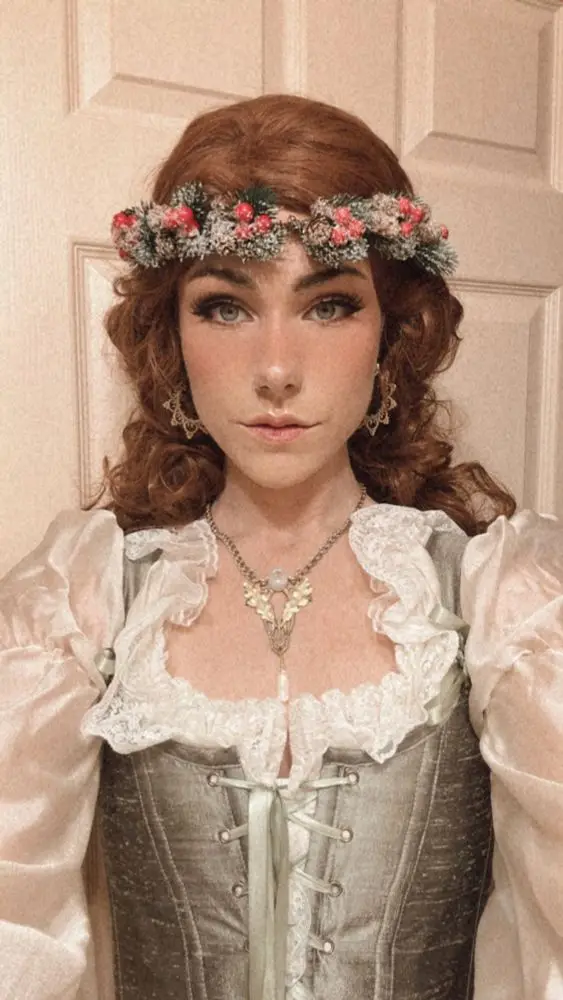A woman with red hair and a floral crown wears a historical-style outfit with a corset and lace collar, looking directly at…