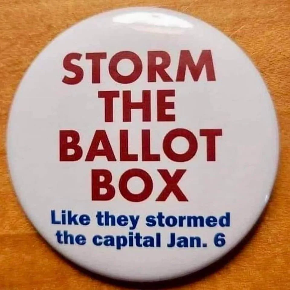 A white circular button with red text reading 'STORM THE BALLOT BOX' and smaller blue text below saying 'Like they stormed…