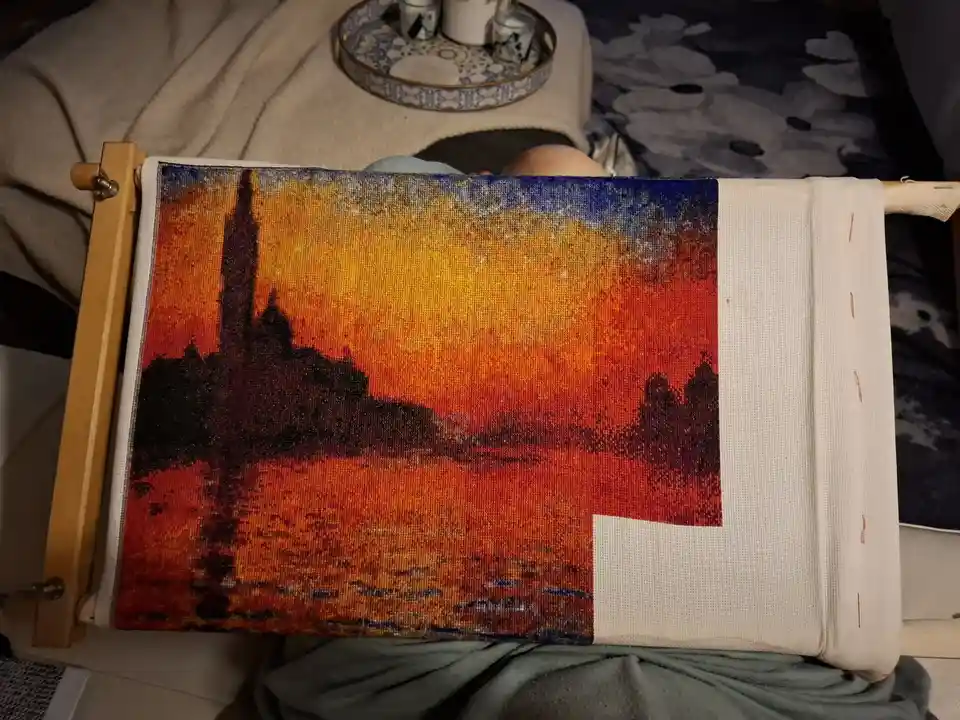 A partially completed cross-stitch artwork depicting a vibrant sunset over a cityscape with a tall spire, mounted on a…