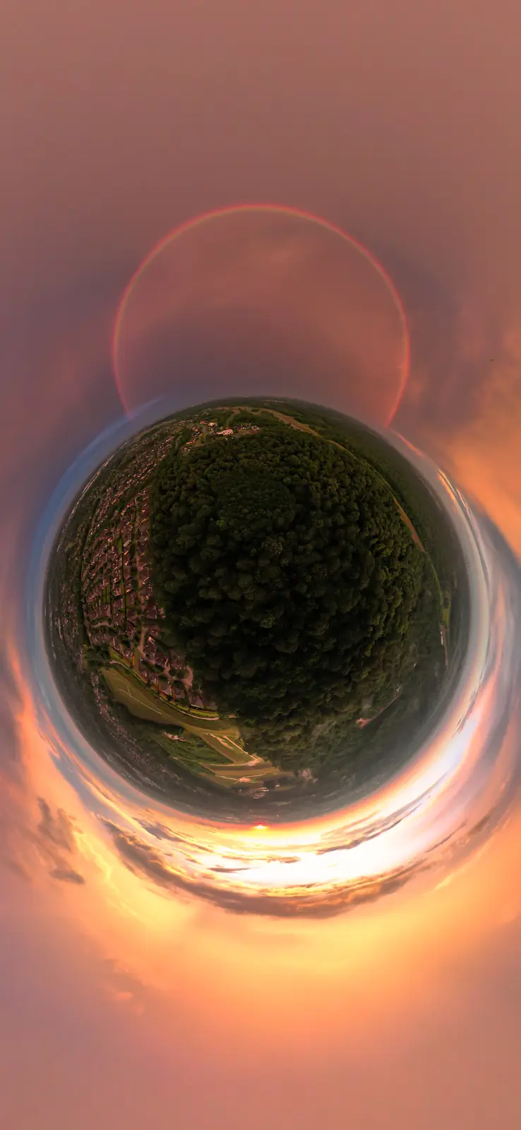 A 360-degree fisheye panorama of a sunset over a forest and suburban area, distorted into a spherical 'little planet' effect…