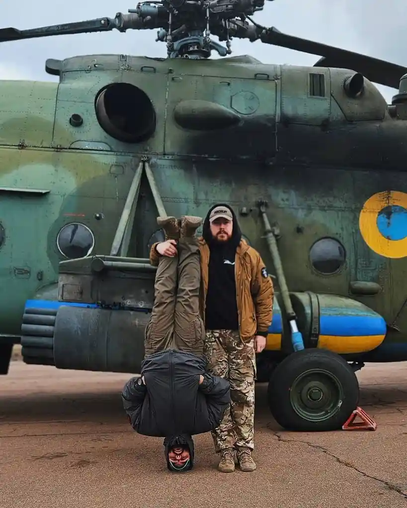 A man in a tan jacket and camouflage pants stands in front of a large green military helicopter with Ukrainian insignia…