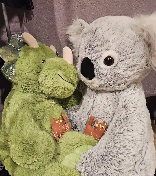 A green plush dinosaur with horns and a smiling face is hugging a large grey plush koala. Both stuffed animals have small…