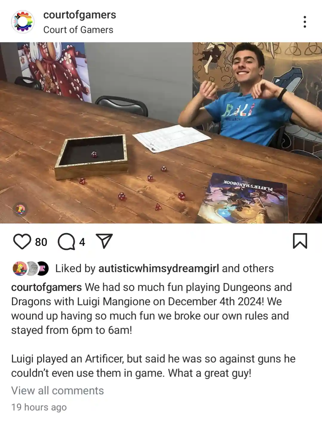 A young man in a blue t-shirt sits at a wooden table with a triumphant, smug expression, making a 'victory' gesture with…