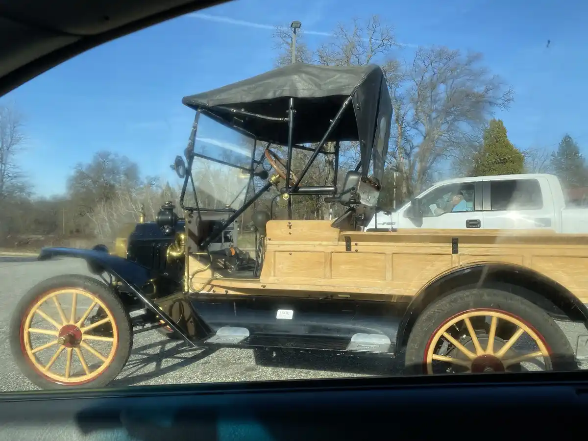 A photo taken from inside a car shows a vintage black and wood-paneled automobile with brass accents and spoked wheels…
