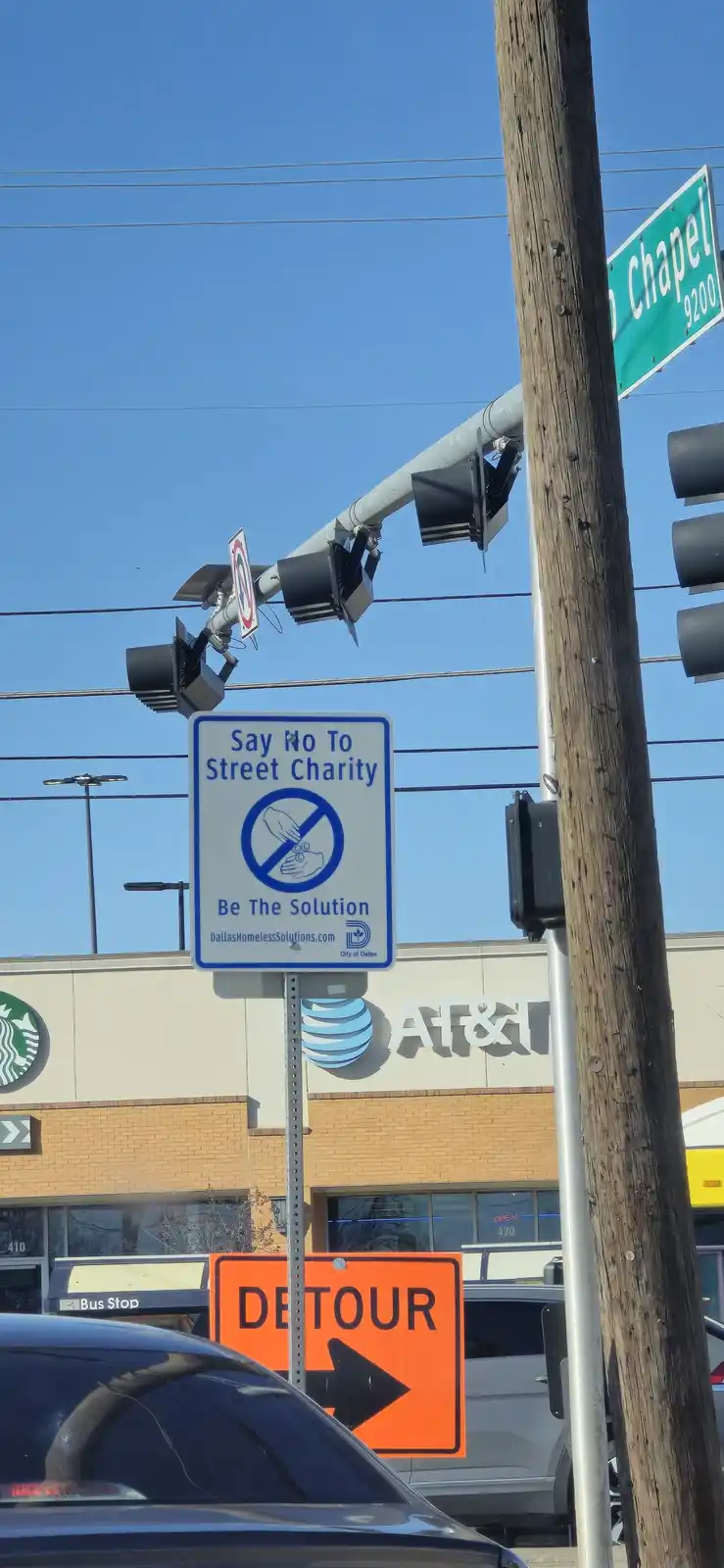 A street sign in Dallas reads 'Say No To Street Charity' with a symbol of a hand giving coins crossed out, followed by 'Be…