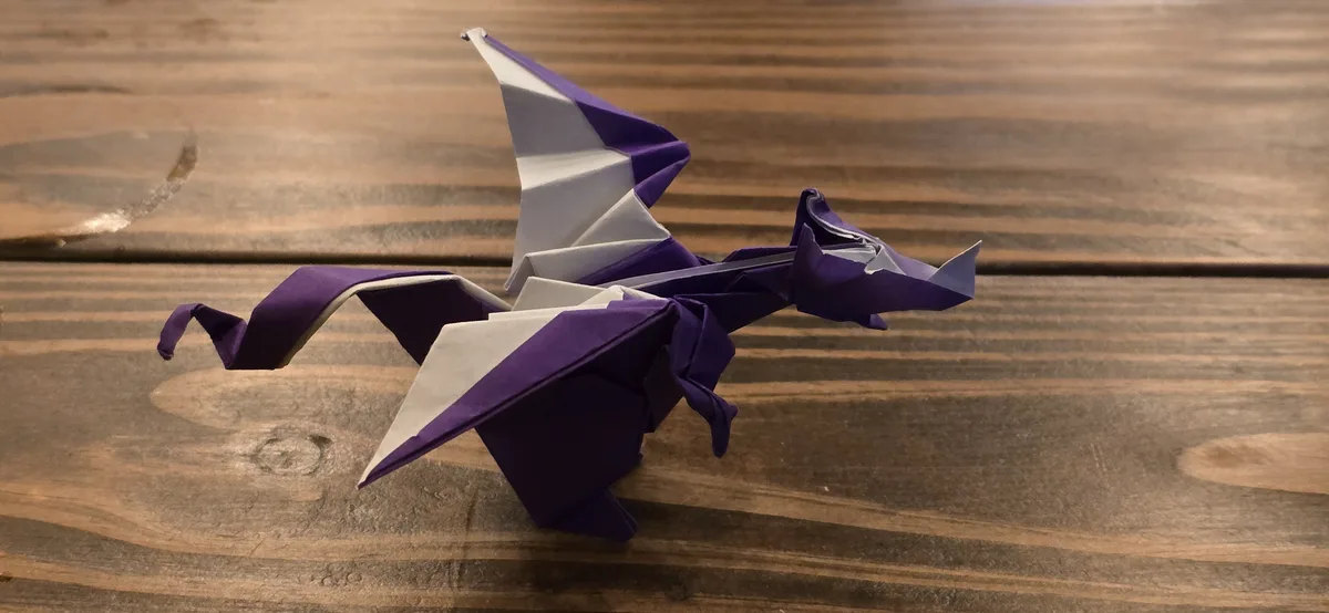 A purple and white origami dragon sits on a wooden surface, its paper folds forming wings, tail, and head. The overlaid text…
