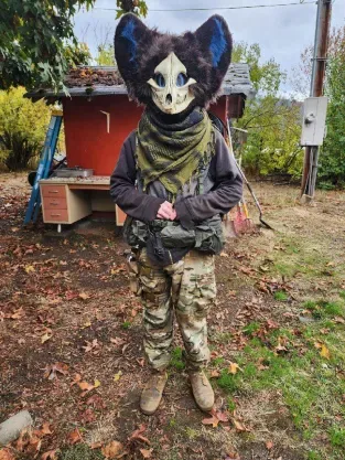 A person stands outdoors in a yard wearing a full-body camouflage outfit and a large, furry, skull-faced cat mask with blue…