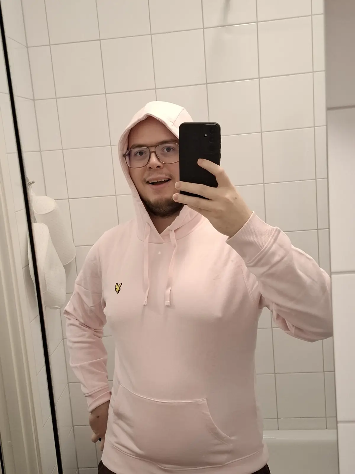 A man with glasses and a light pink hoodie takes a mirror selfie in a tiled bathroom, holding a black phone. He has a…
