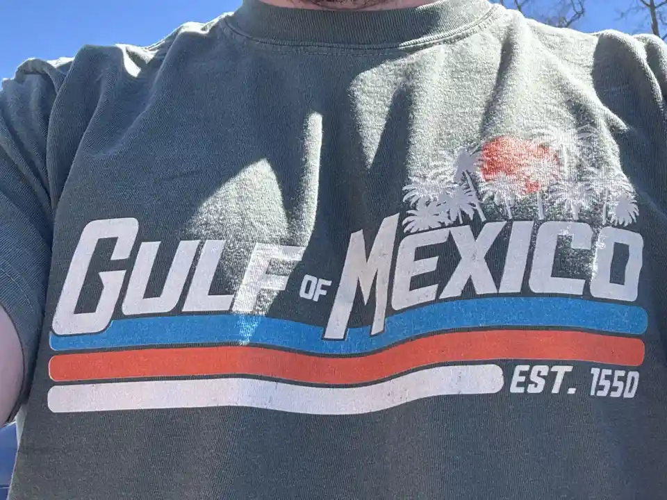 A close-up of a person wearing a faded gray t-shirt with a graphic that reads 'GULF OF MEXICO' in large white letters, with…