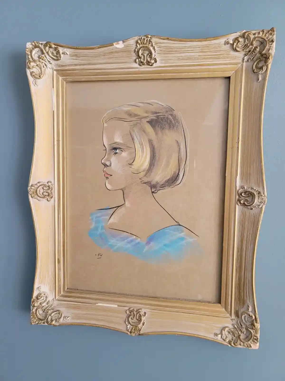 A framed pastel drawing of a young girl in profile, wearing a blue top, hangs on a blue wall. The ornate gold and white…