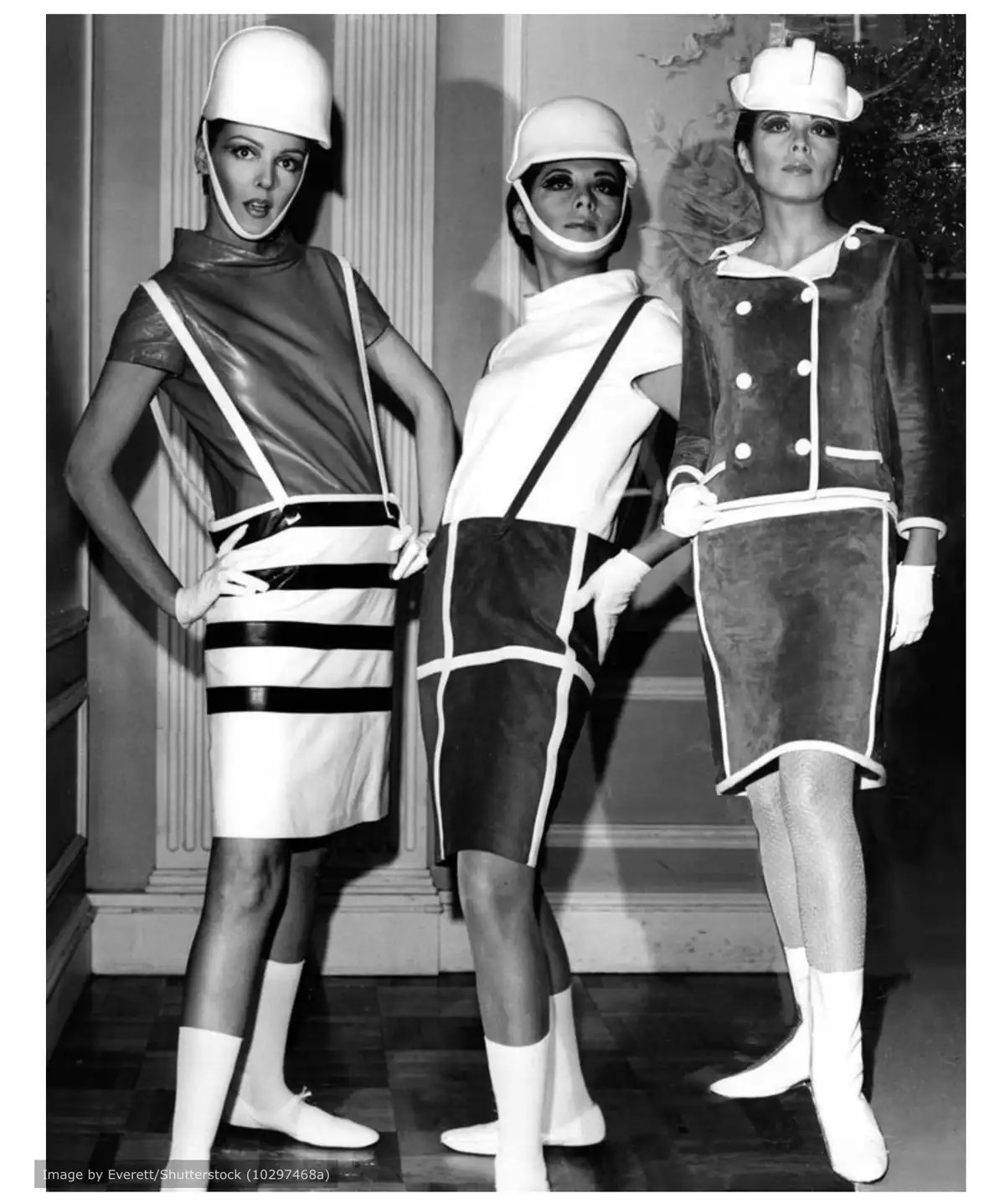 Three women in 1960s futuristic fashion, wearing white helmets and geometrically patterned outfits, pose with one looking…