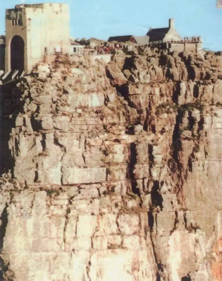 A vintage photograph shows a steep, rocky cliffside with buildings perched at the top, including a prominent stone tower and…
