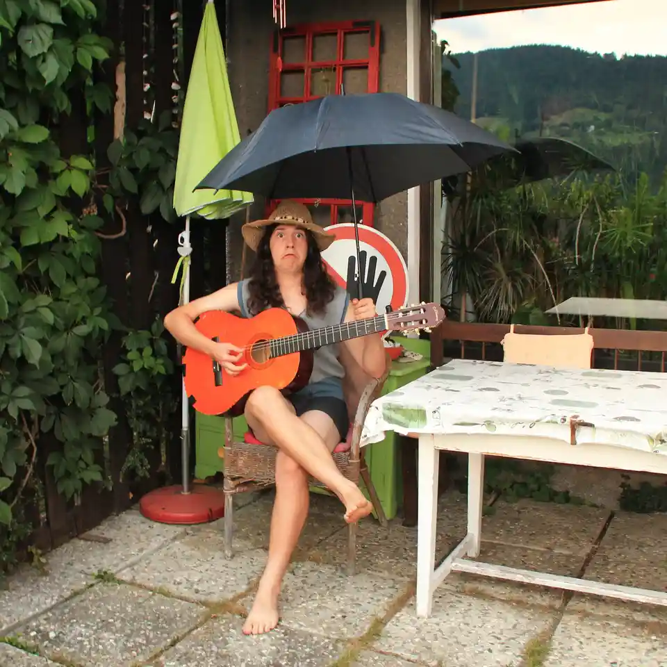 A person with long dark hair wearing a straw hat sits barefoot on a wicker chair outdoors, holding a bright orange acoustic…
