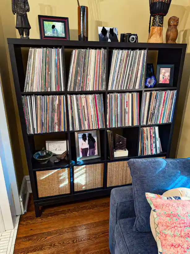 A black cube-style bookshelf is filled with vinyl records standing upright in rows, with some framed photos and decorative…