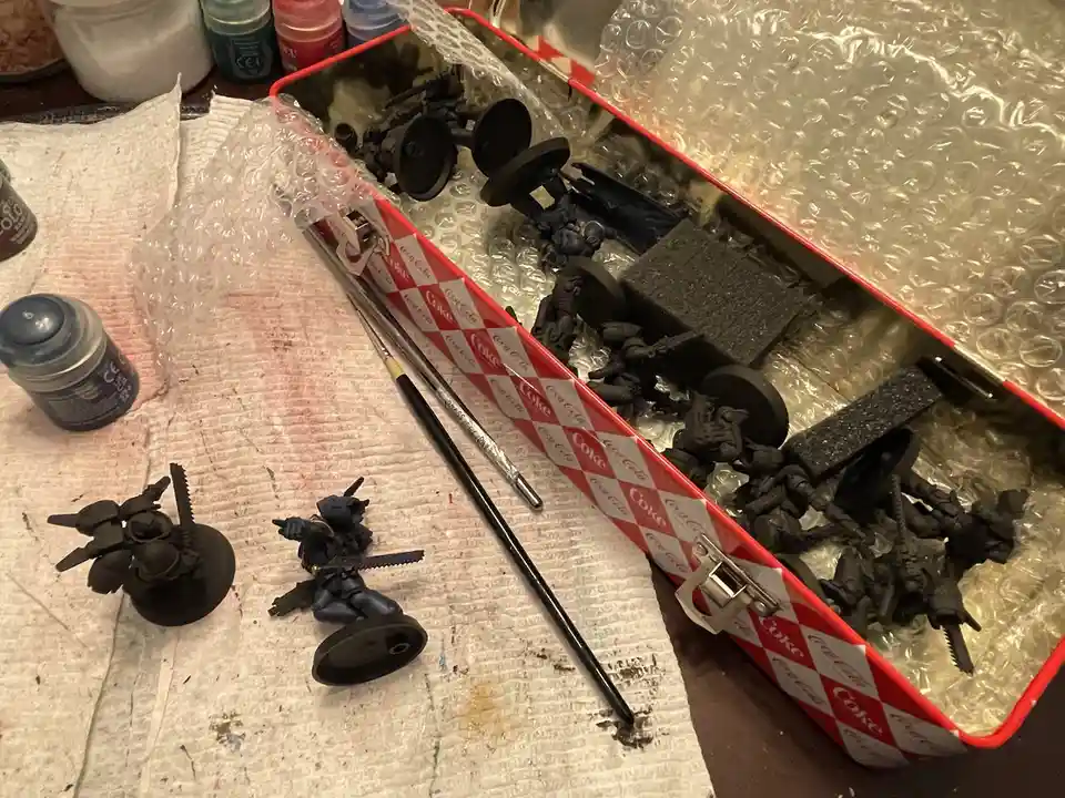 An open red and white Coca-Cola branded metal tin filled with unpainted black miniature figurines and bubble wrap, sitting…