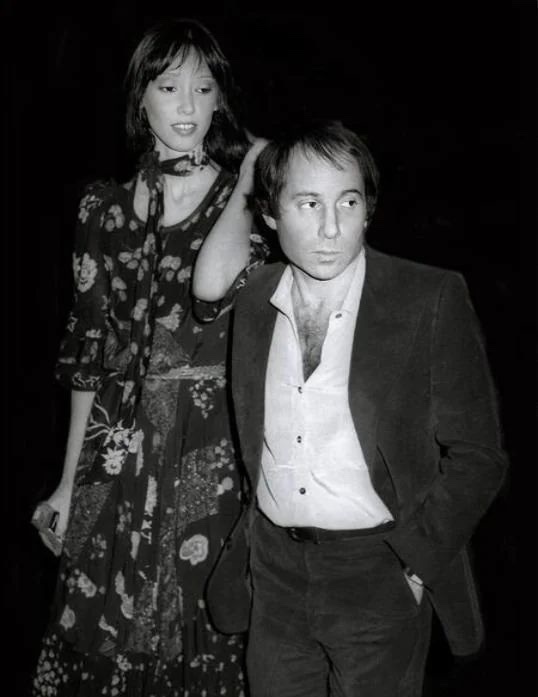 A black-and-white photo shows a woman in a floral dress standing behind a man in a suit, her hand on his head. The man has a…