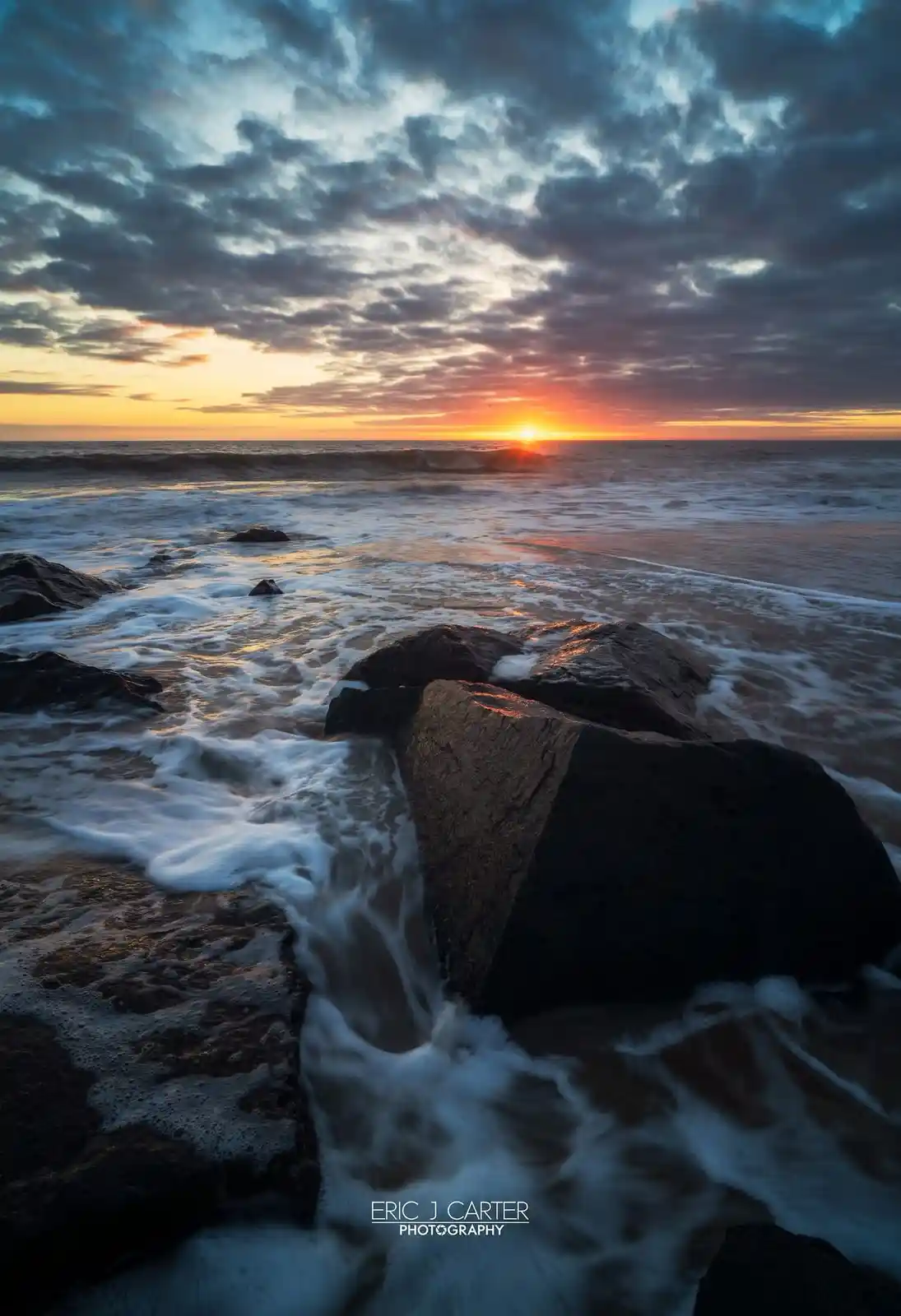 A long-exposure photograph of a dramatic ocean sunset with waves crashing over dark rocks, the sky filled with textured…