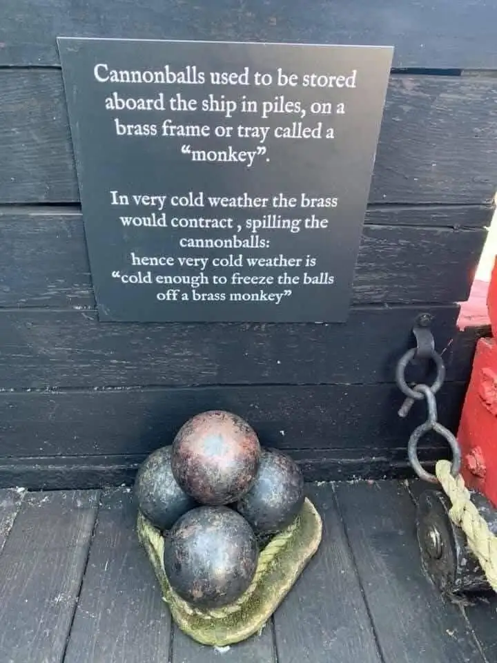 A dark wooden sign explains the origin of the idiom 'cold enough to freeze the balls off a brass monkey,' describing how…