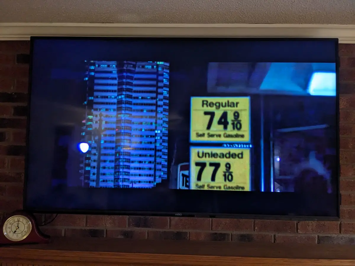 A television screen mounted on a brick wall displays a nighttime scene with illuminated skyscrapers and a gas station price…