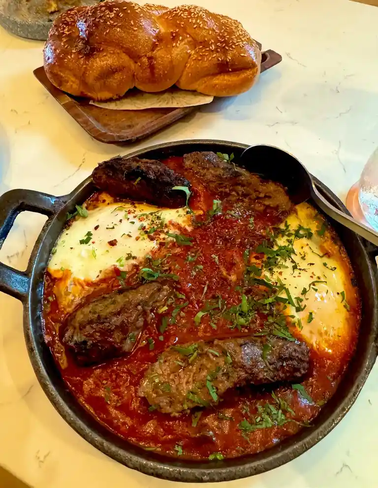 A close-up photo shows a cast iron skillet filled with a rich red tomato sauce, topped with melted cheese, chopped green…