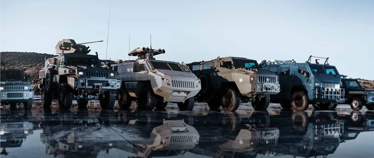 A row of heavily armored military vehicles, including Humvees and other tactical trucks, are parked in a shallow puddle that…