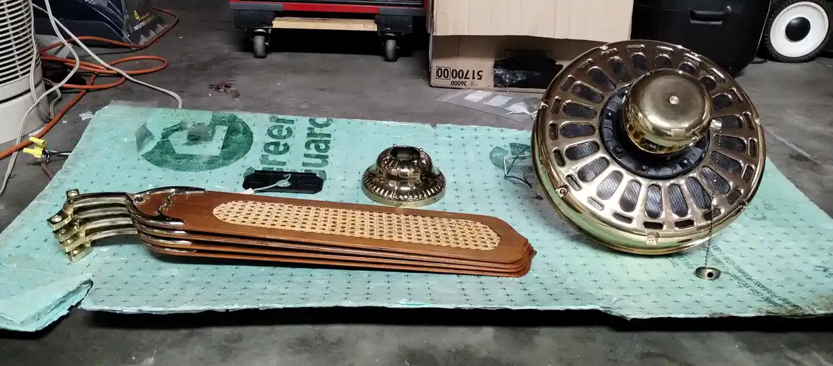A disassembled, ornate ceiling fan with brass finish and wooden blades lies on bubble wrap in a garage. The fan's motor…