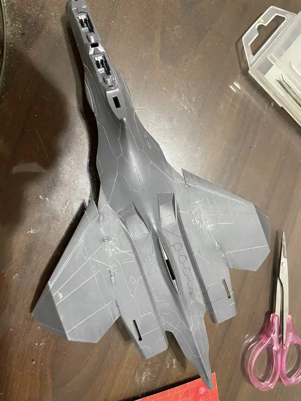 A gray, unfinished plastic model of a futuristic fighter jet sits on a wooden workbench, surrounded by modeling tools…