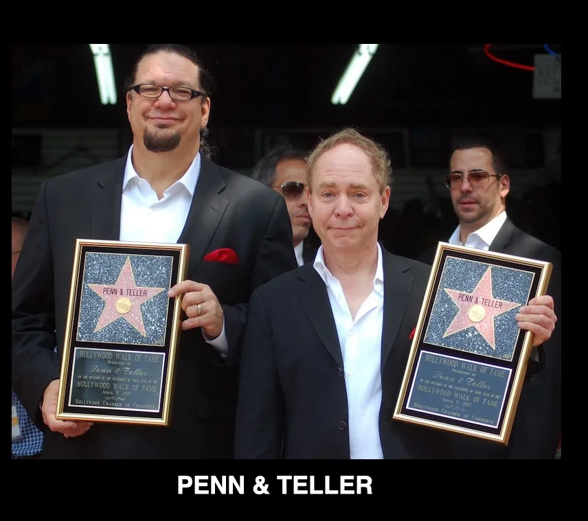 Two men in suits hold identical framed replicas of their Hollywood Walk of Fame star plaques, smiling for the camera. The…